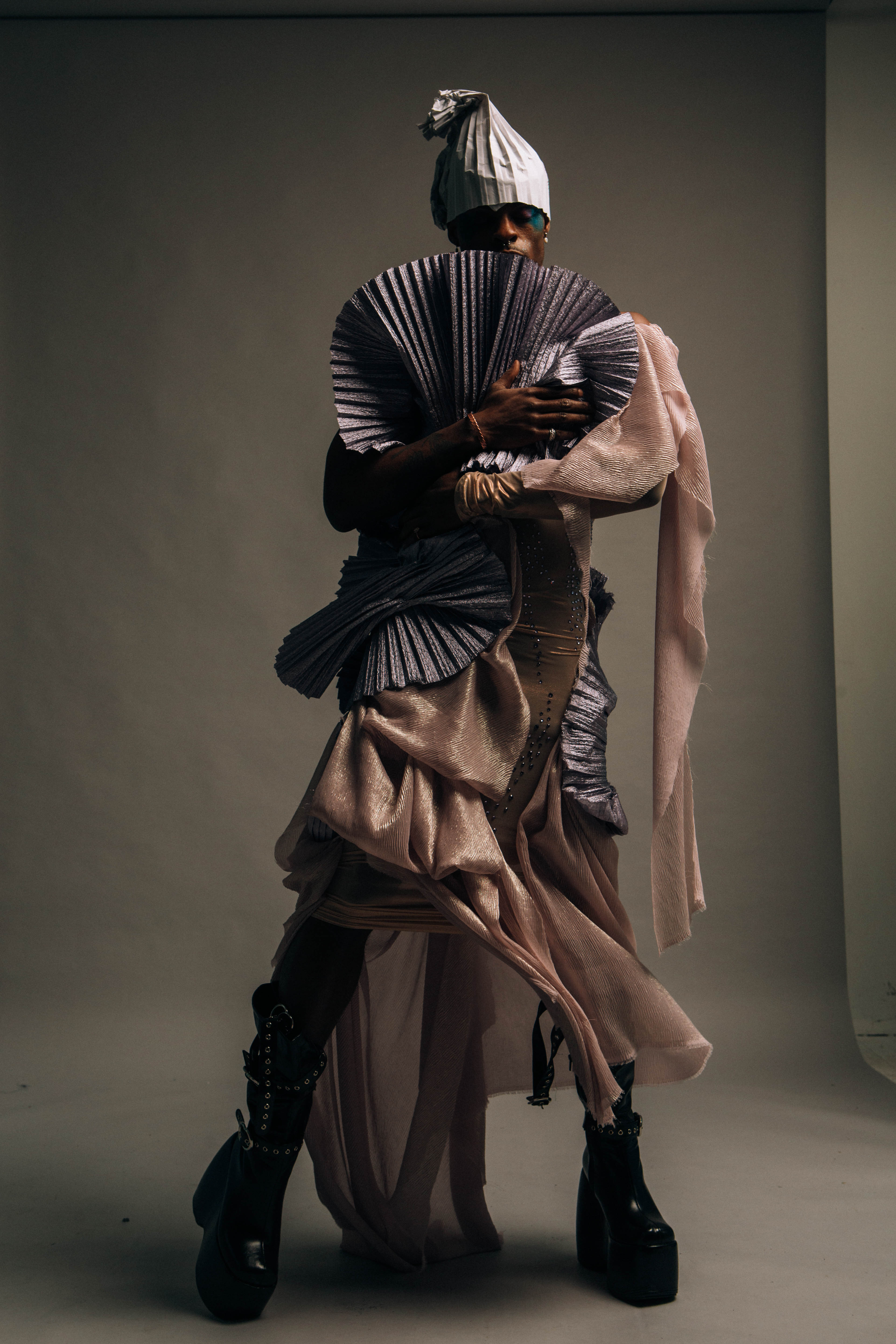 The same model and outfit from the previous photo is displayed in a different pose, showing another angle to the garment's draping; the model holds the chest-level purple pleated fabric like a fan. The background is dark grey.