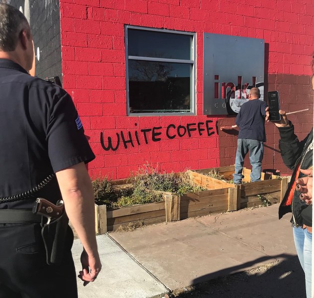 A person paints over graffiti "white coffee" painted on side of a red building while cop watches.