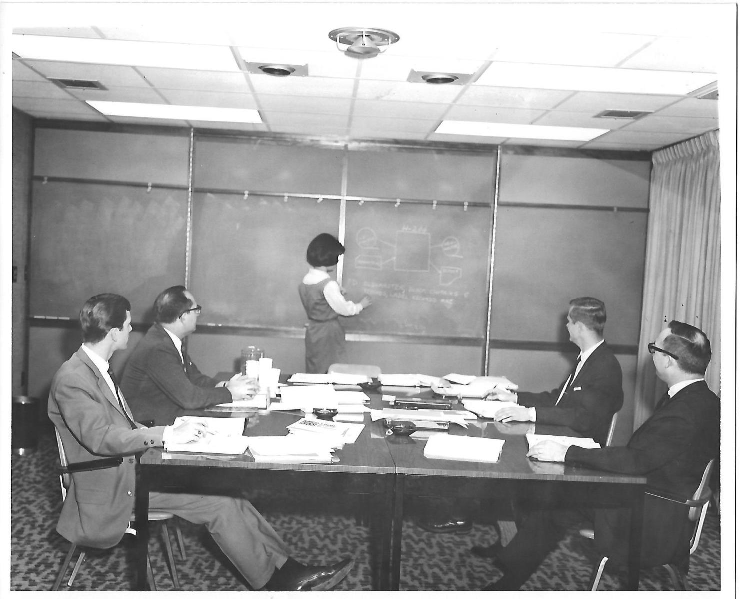 A black and white photo of four men in suits and a woman in a dress. The woman is drawing diagrams on the blackboard, while the men watch from a table filled with papers.