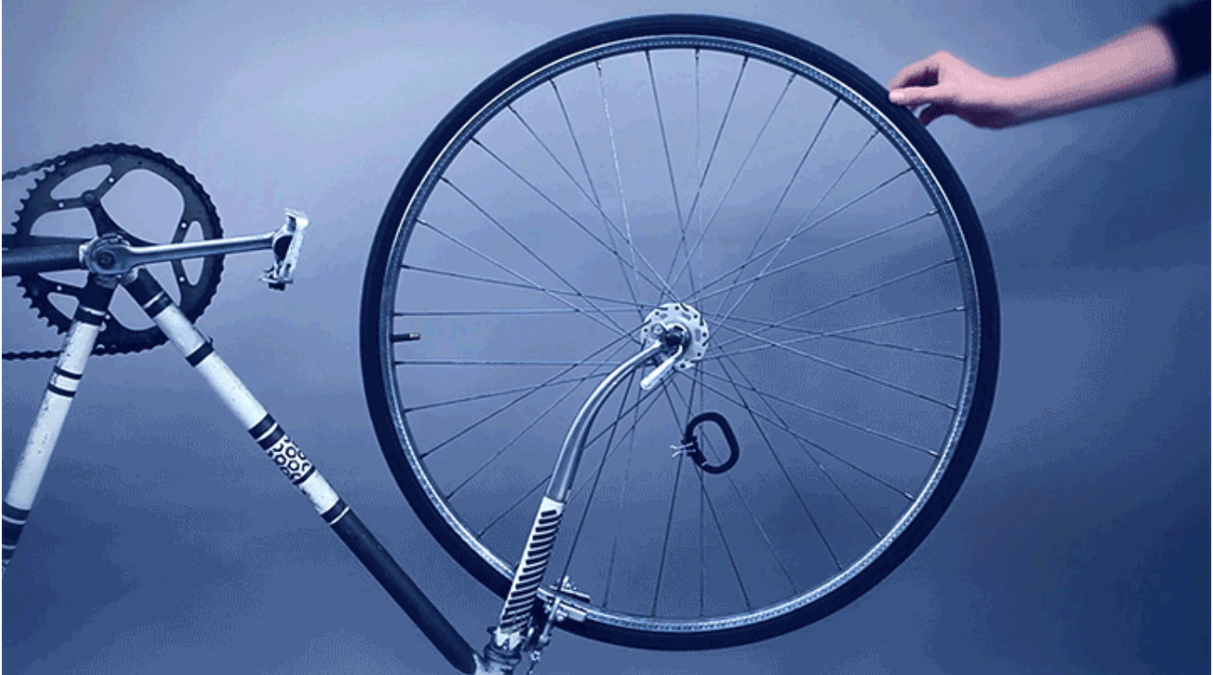 A hand turns the wheel of an upside down bicycle, with a Fitbit attached to one of the spokes.