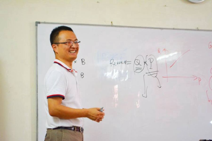 A man smiling in front of a whiteboard, holding dry erase markers.