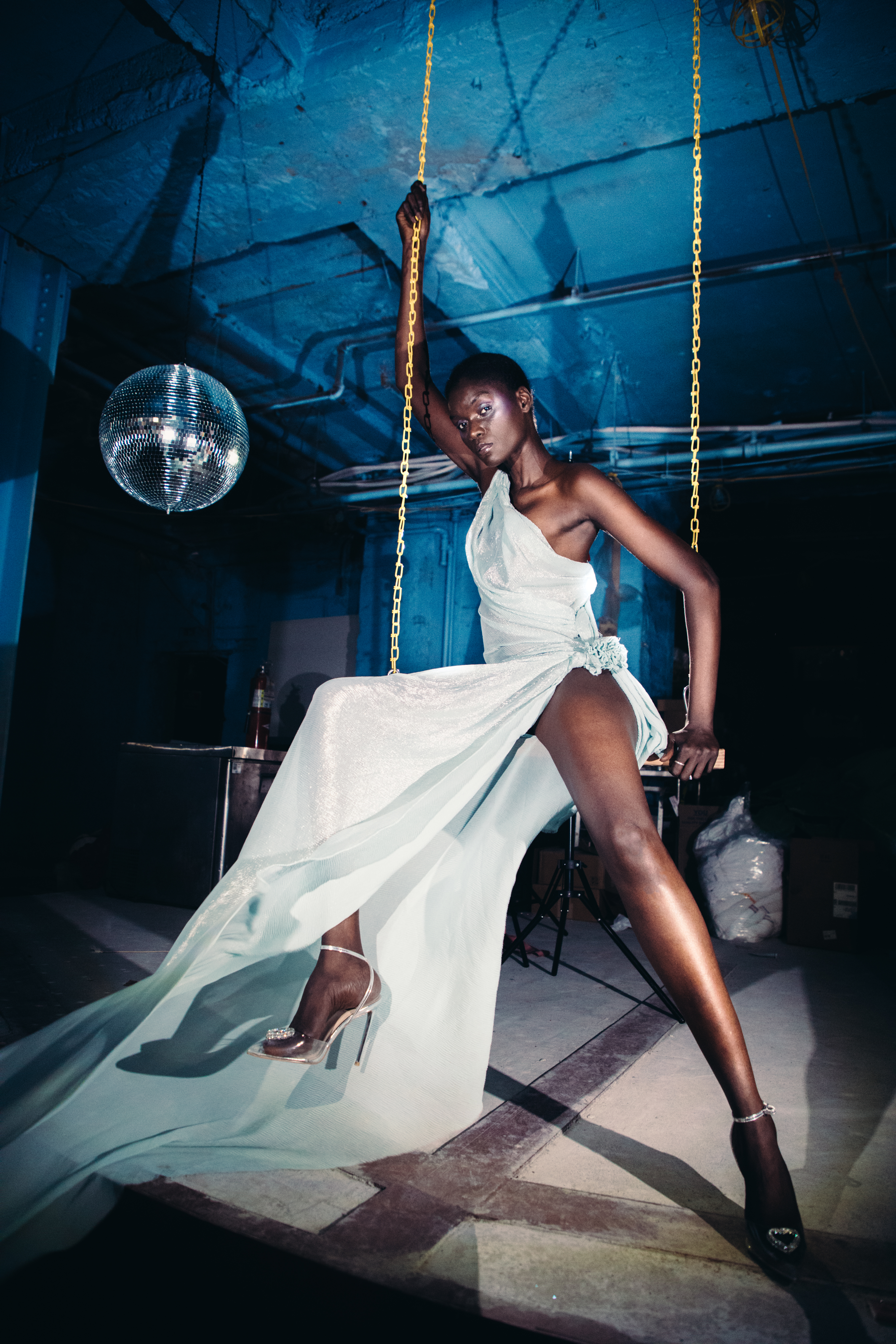 A Black model spins in a black and white pleated head wrap, with a blue, white,
     and silver-grey pleated strapless shirt tied with some ribbons. Translucent light
     green-blue fabric fans out to the model's right. The skirt of their outfit is made
     of a shimmery light green fabric with smaller pleats, and it drapes across their
     legs, against a white backdrop.