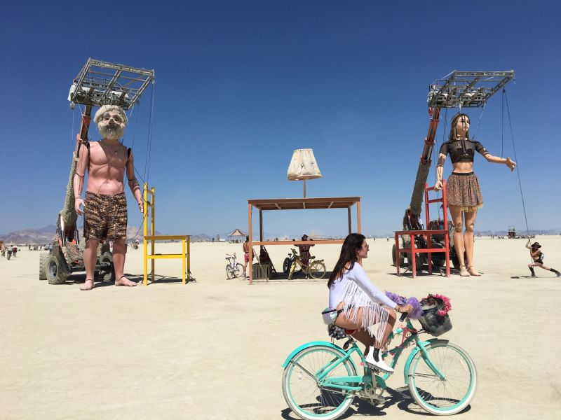 A person biking across a dessert on a teal cruiser bike, with gigantic human puppets behind.