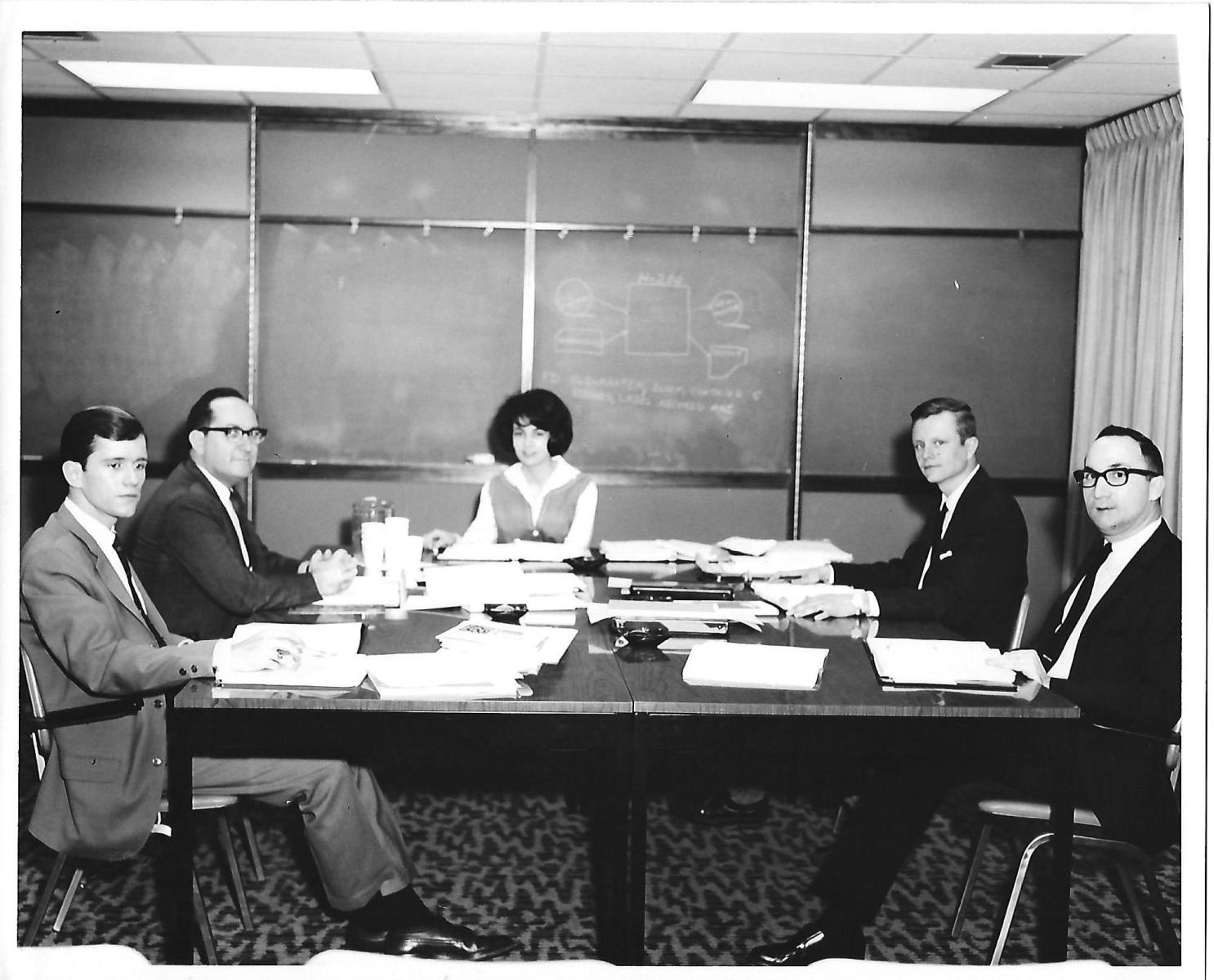 A black and white photo of four men and one woman at a paper-filled desk with a chalkboard with diagrams in the background. The men wear suits and the woman is wearing a white shirt with a vest, possibly a dress.