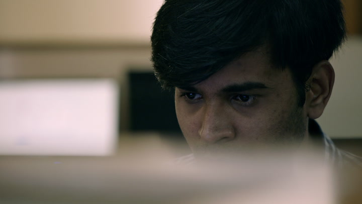 A man of Indian descent looking over the top of a computer screen.