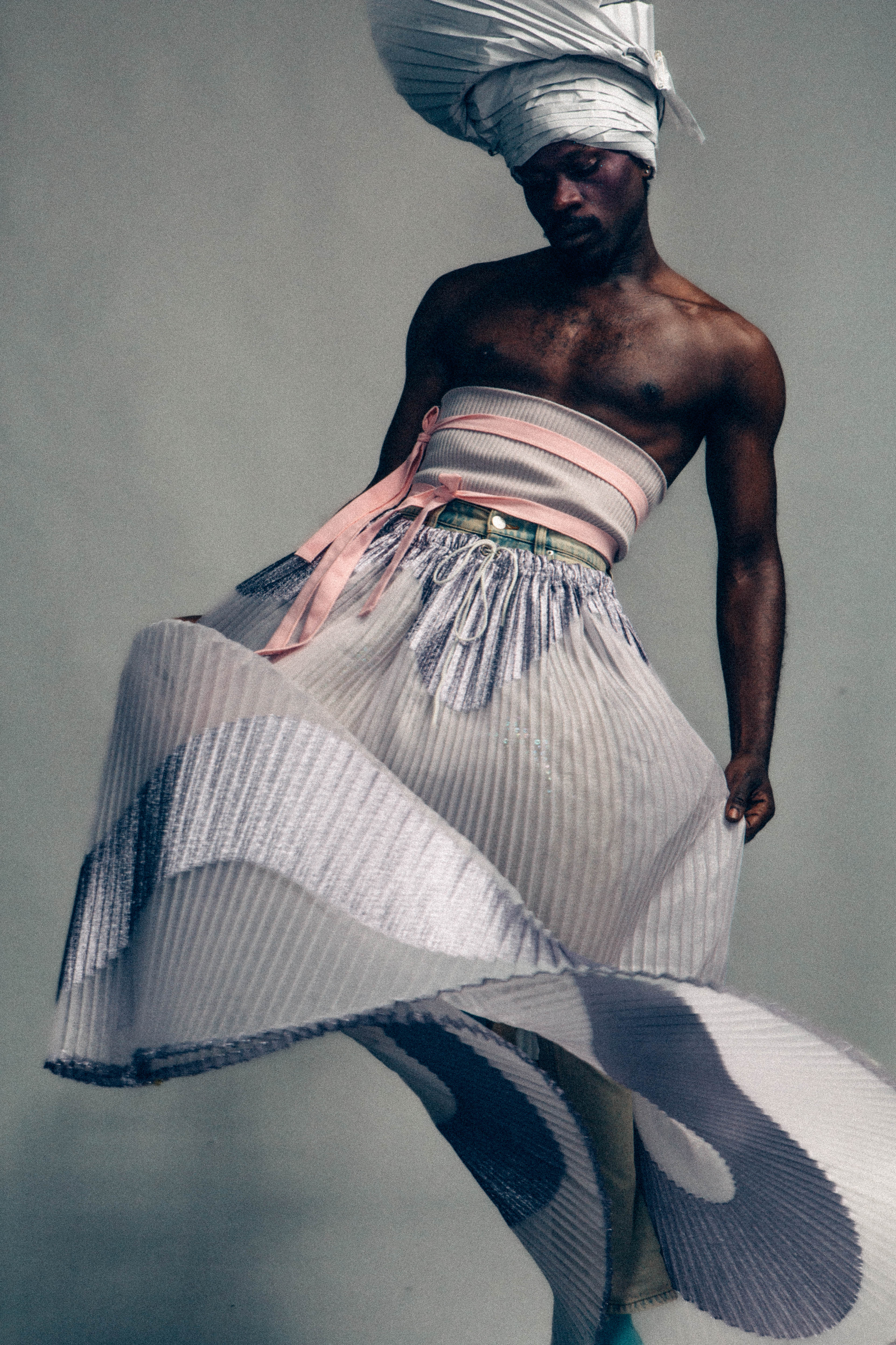 Against a grey background, a Black model poses with a light blue pleated head wrap, a ribbon-tie blue and pink tube top, a glimpse of a denim skirt underneath an elasticated maxi skirt swishing up, in pleated shimmery and matte purple fabric. They hold their skirt at both sides as it twirls around them.