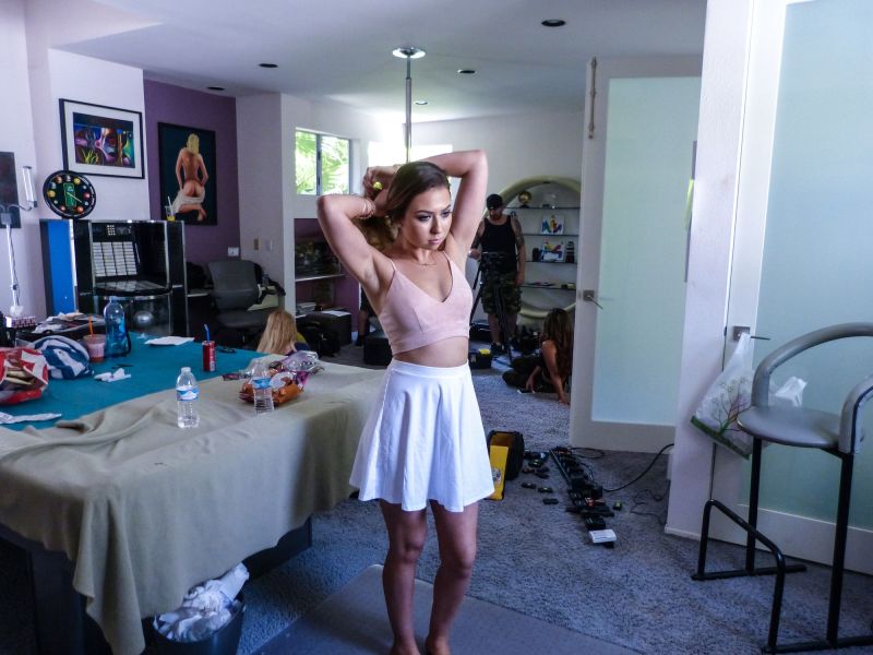 A woman in a crop top and white skirt stands in front of a bed on a porn set, water bottles, equipment, and other workers behind her.