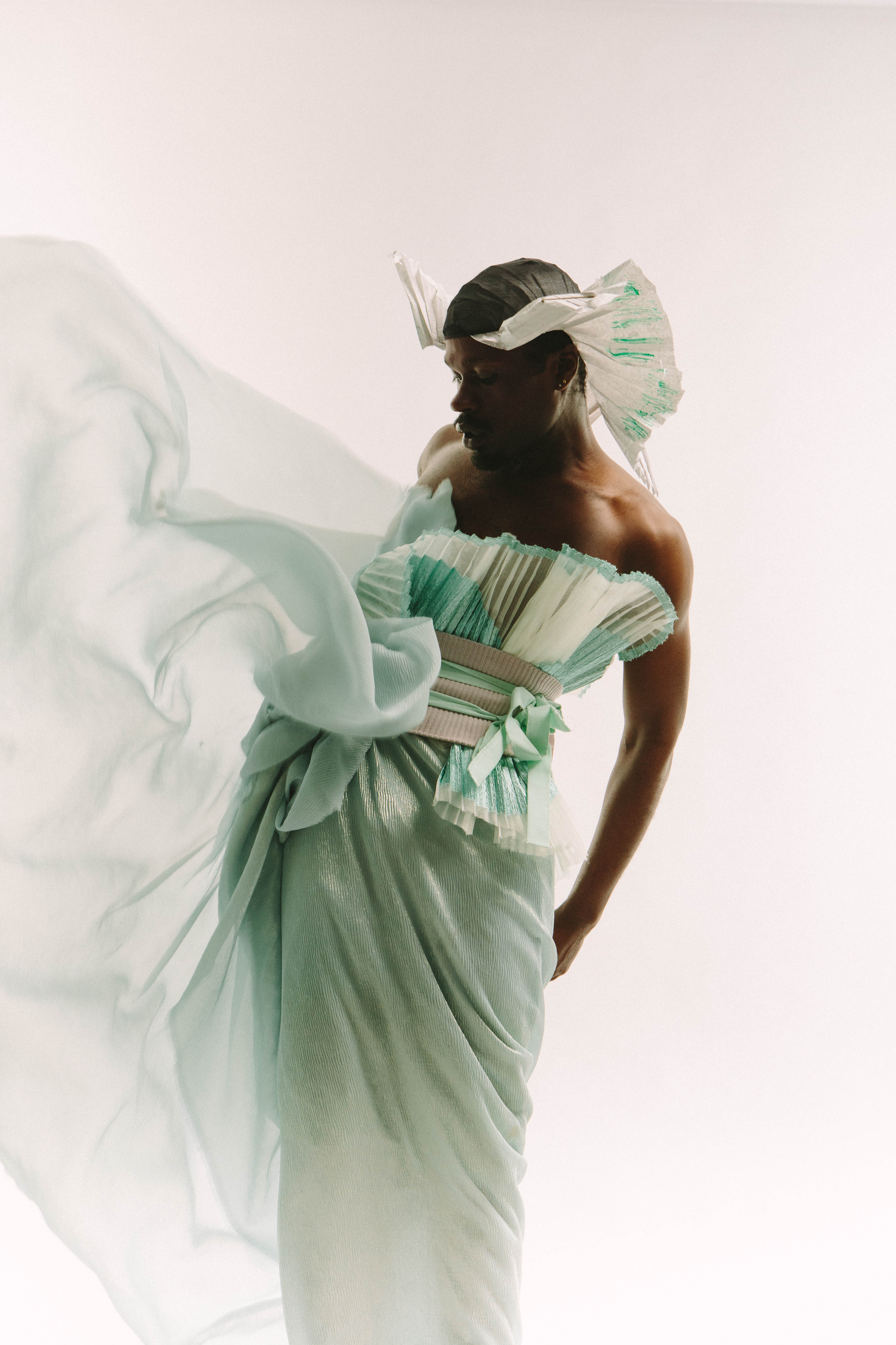 A Black model spins in a black and white pleated head wrap, with a blue, white, and silver-grey pleated strapless shirt tied with some ribbons. Translucent light green-blue fabric fans out to the model's right. The skirt of their outfit is made of a shimmery light green fabric with smaller pleats, and it drapes across their legs, against a white backdrop.