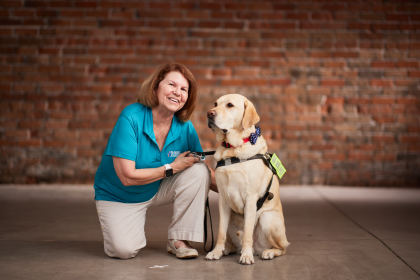 Portrait of Teresa Galgano wearing a teal BVA polo, kneeling next to her guide dog, Donald, who is wearing a harness and American-flag printed bow tie.