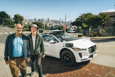 Steve and Newton, two Curry Senior Center community members, walk toward the camera laughing after exiting a Waymo autonomous vehicle