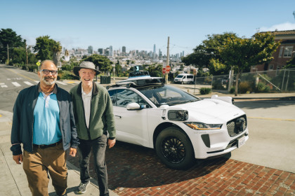 Steve and Newton, two Curry Senior Center community members, walk toward the camera laughing after exiting a Waymo autonomous vehicle