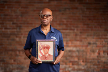 Portrait of Sheldon McKinney wearing a blue BVA polo shirt, standing in front of a brick wall holding a framed photograph of his younger self.