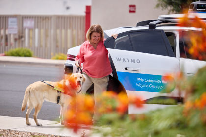 Teresa and her guide dog, Donald, stepping out of a Waymo vehicle onto the sidewalk in front of a bush with orange flowers. The door to the Waymo is printed with the text "Ride today with the Waymo One app" on a blue stripe.