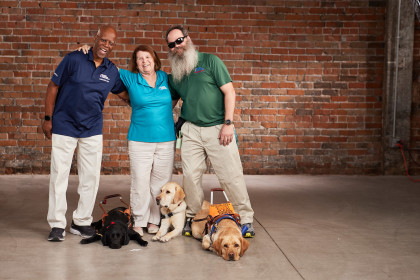 Three Blinded Veterans Association members, Sheldon, Teresa, and Rob, stand wearing BVA polo shirts and khaki pants with their arms around each other, and their guide dogs at their feet.