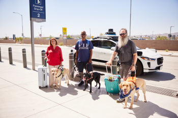 Three Blinded Veterans Association members standing with their guide dogs in front of a Waymo at the Phoenix Sky Harbor Airport
