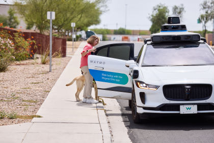 Woman and her guide dog standing on the sidewalk, opening the door to an autonomous Waymo vehicle. The door of the vehicle has a blue decal that reads "Ride today with the Waymo One app" in white text.