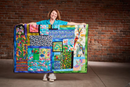 Teresa smiling and standing in front of a brick wall, holding up a large handmade quilt