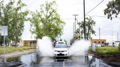 A white, Waymo Pacifica minivan splashing through a puddle at a closed-course testing facility