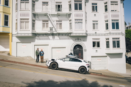 Newton and Steve standing on a sloped sidewalk in front of a white building, as a white Waymo autonomous vehicle pulls up in front of them