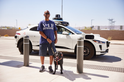 Sheldon and his guide dog, Nora, both stand smiling toward the camera in front of a Waymo autonomous vehicle.