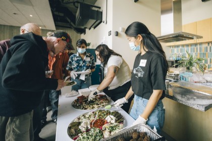 Volunteers stand in a kitchen, serving Mediterranean food to a line of older men 