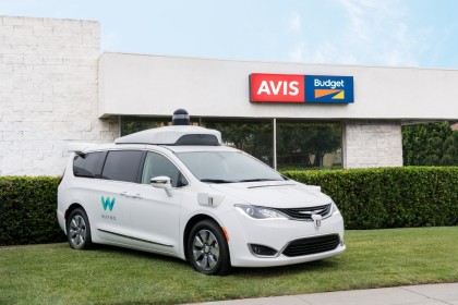 A photo of a white, Waymo Pacifica minivan in front of an Avis office