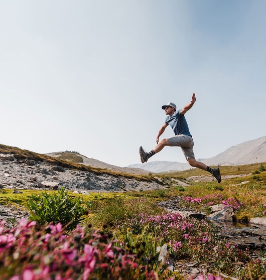A hiker leaps across a small stream in an alpine valley amid wildflowers.
