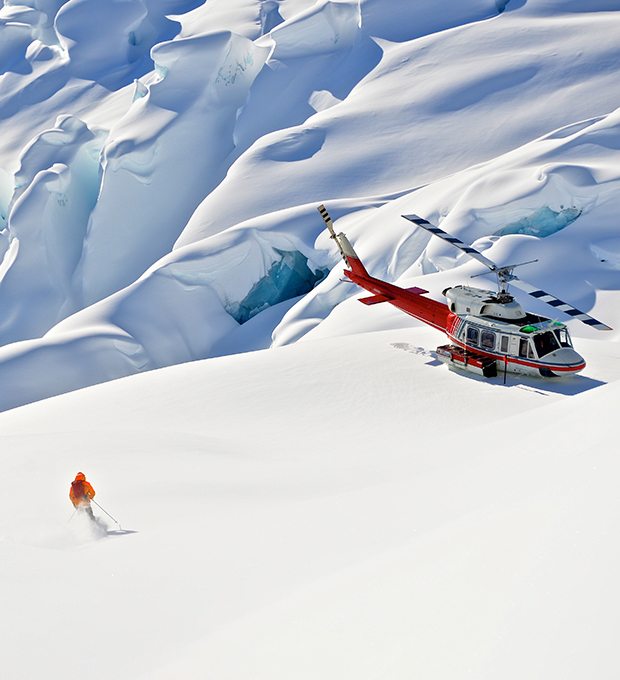 A skier carves turns in fresh powder as he returns to a helicopter perched on a mountain ridge.