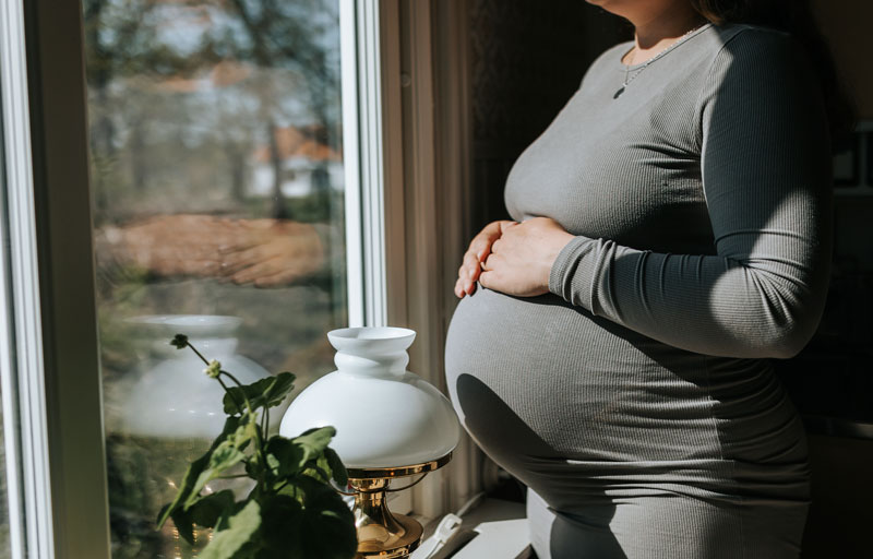 Gravid kvinna står i solen framför fönstret, med händerna på magen. Hon har en grå långärmad klänning på sig.