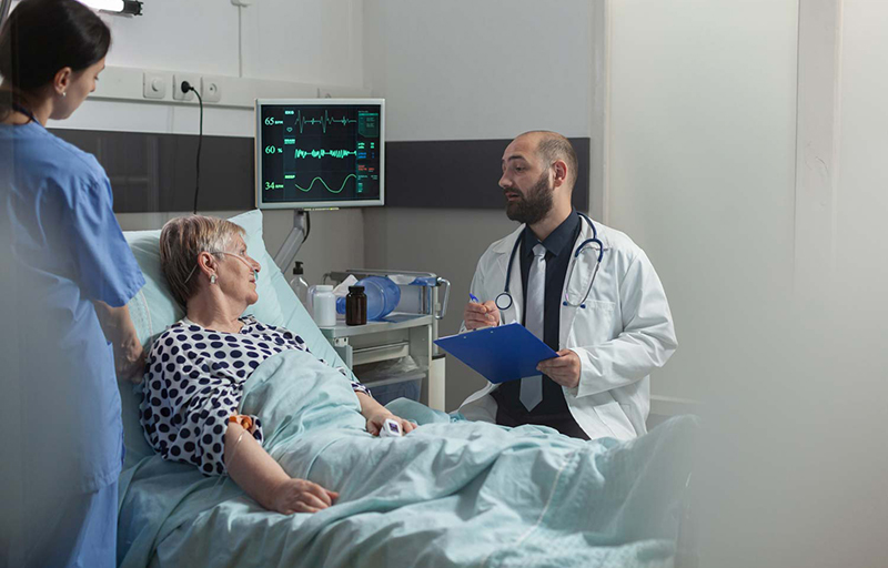 Patient ligger i en sjukhussäng med en doktor och en sköterska bredvid sig.