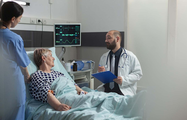 Patient ligger i sjukhussäng och två läkare står bredvid
