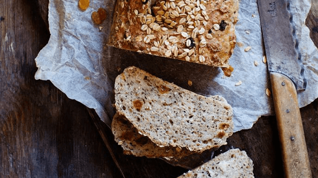 Brödlimpa med frön och torkad frukt, uppskuren på ett bord med brödkniv bredvid.