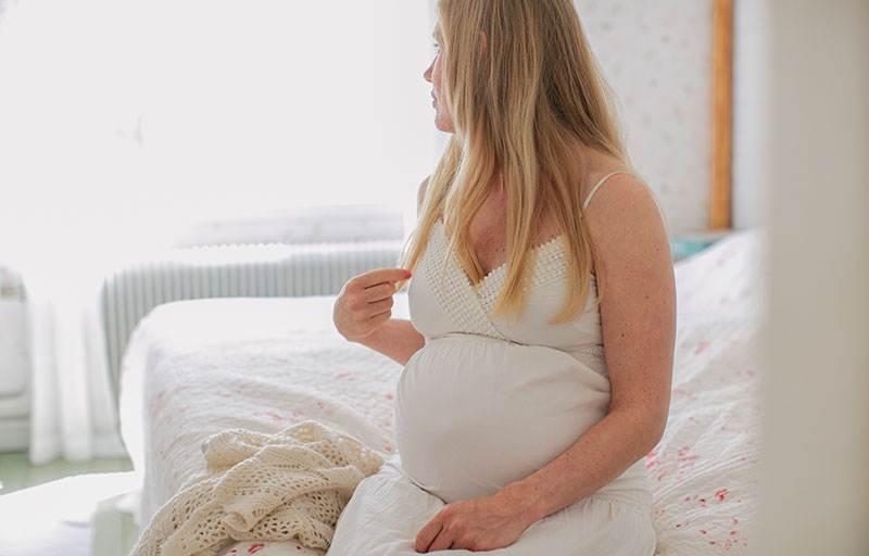 Gravid kvinna tittar bort från kameran i vit klänning. Hon sitter i sängen.