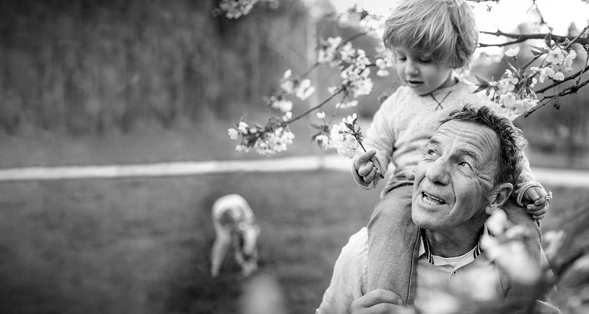 Barnbarn sitter på farfars axlar, träd i vårblom. 