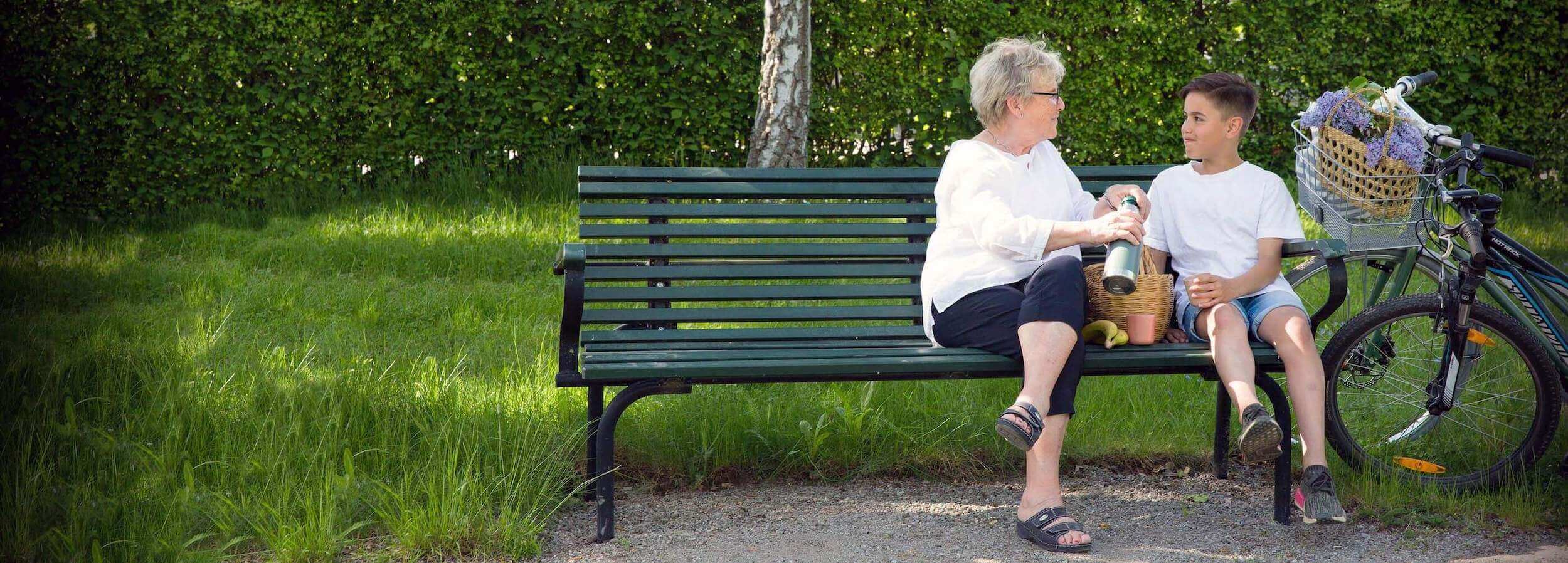 Äldre kvinna och pojke sitter på en parkbänk i somrig park