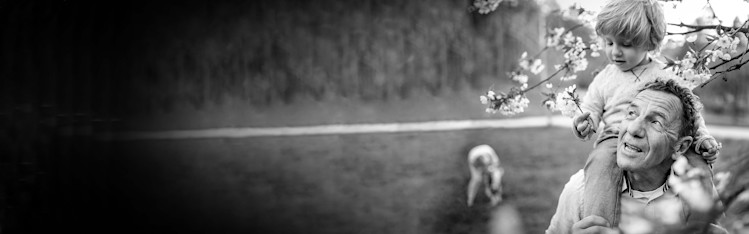 Äldre man med liten pojke på axlarna framför ett träd.