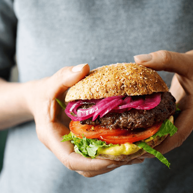 Person håller i en hamburgare. Den är fylld med hamburgerkött, picklad rödlök, currydressing, sallad och tomat.