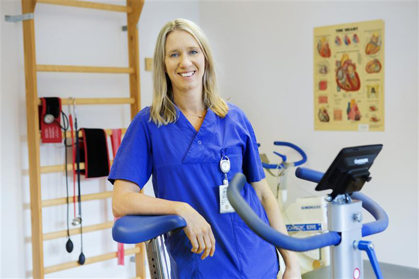 Maria Bäck, forskare, står vid en träningscykel och ler. Hon har en blå skjorta på sig.