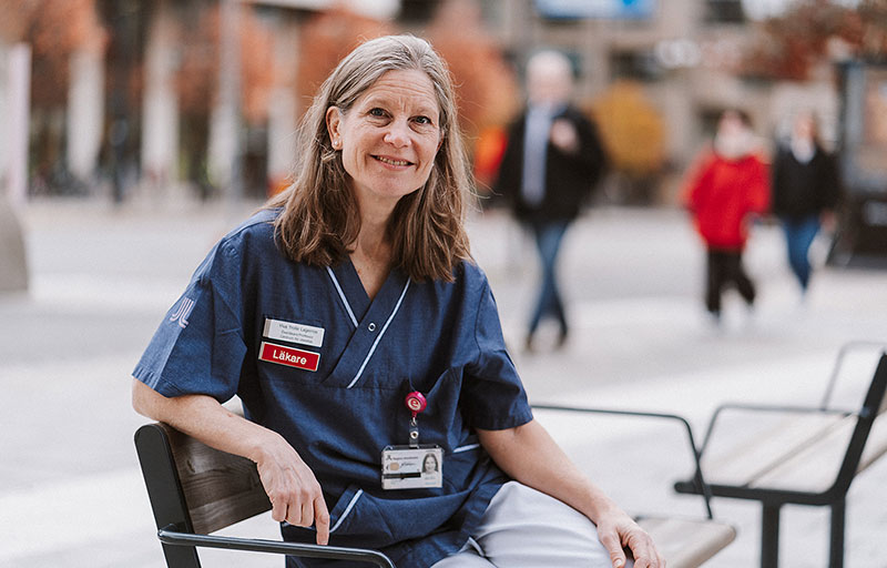 Kvinnlig forskare sitter på en bänk i stan med blå sjukhusskjorta på sig.