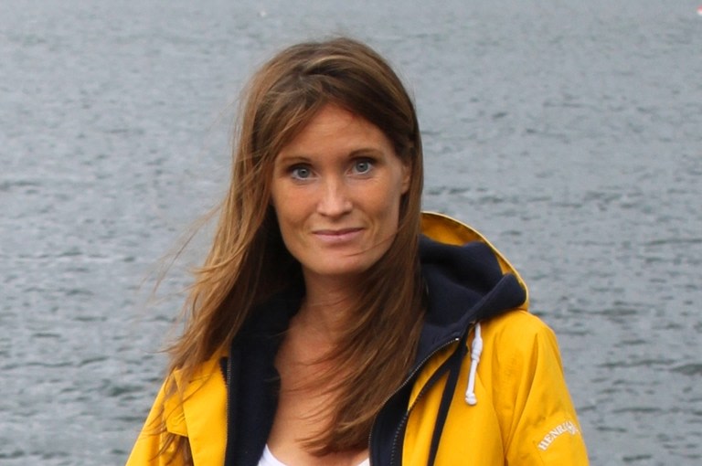 Kvinna med långt brunt hår och gul jacka vid hav