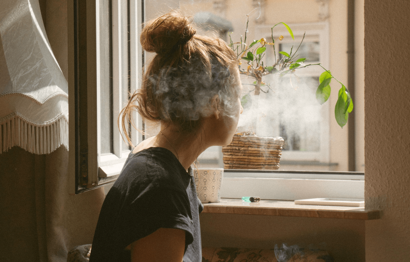 Ung kvinna blåser ut tobaksrök mot ett fönster