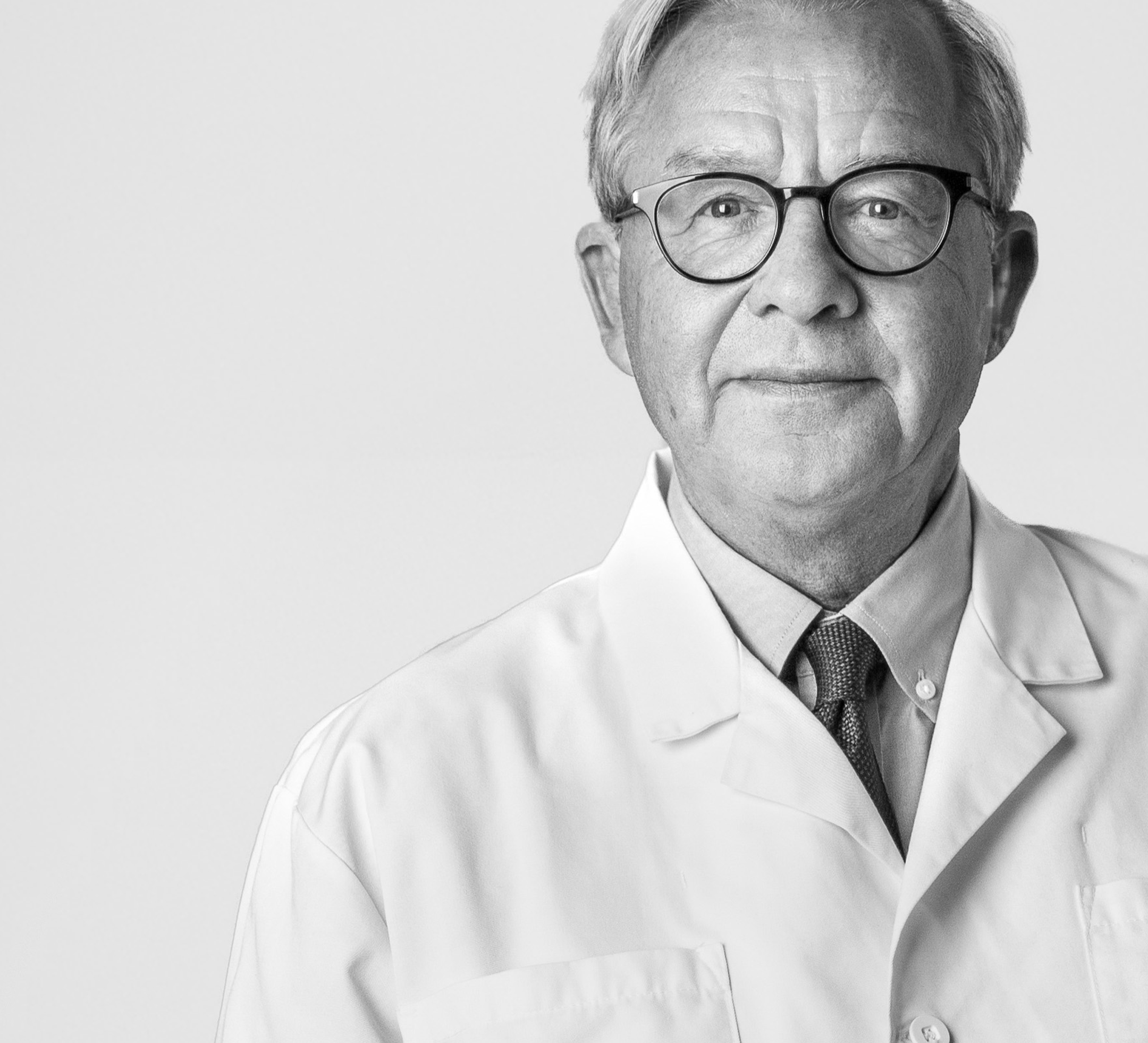 Man i sjukhusrock och skjorta under, tittar in i kameran och ler blygsamt.