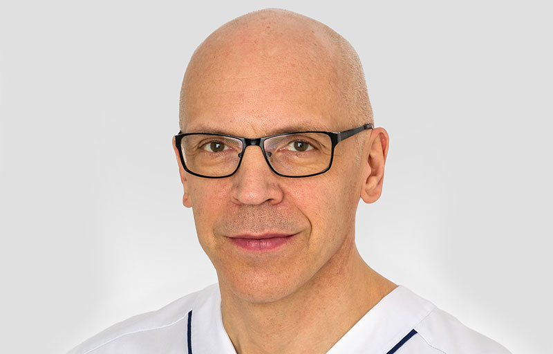 Örjan Friberg ler mot kameran med en grå bakgrund. Han har svarta glasögon på sig.