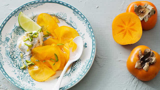 Sharonfrukt med kardemummayoghurt och lime serverade i en skål med lime och pistagenötter