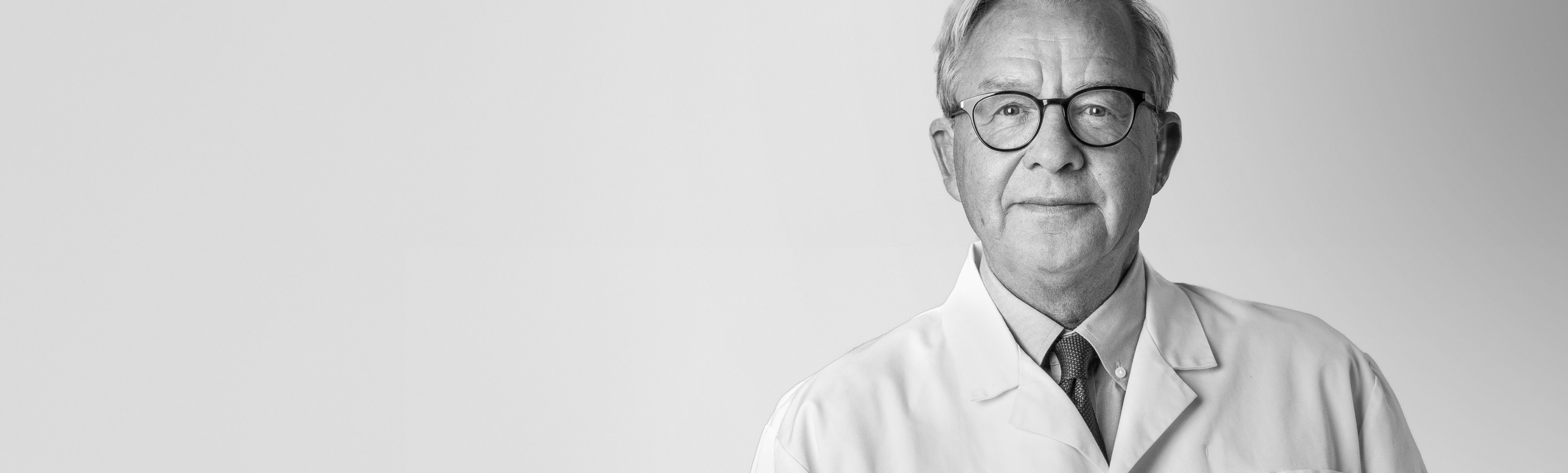 Man i sjukhusrock och skjorta under, tittar in i kameran och ler blygsamt.