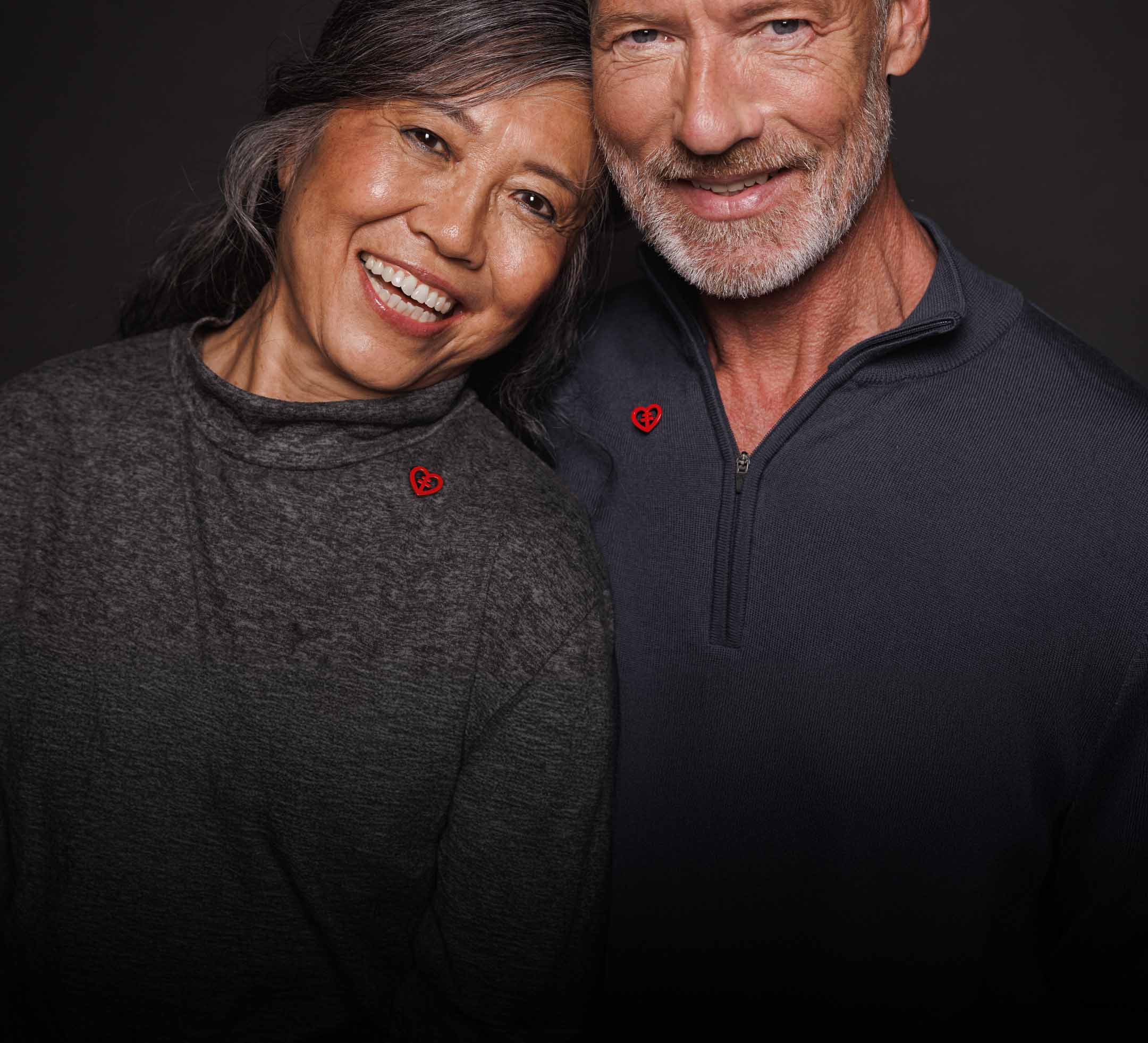 Ett par lutar huvudet mot varandra och ler. De har varsin röd hjärtpin på sina tröjor.