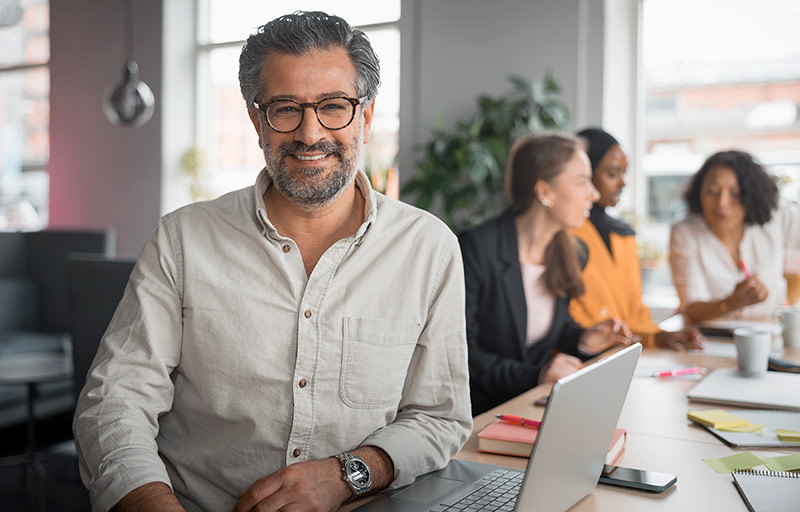 Man med glasögon sitter på kontor bredvid laptop och ler stort.