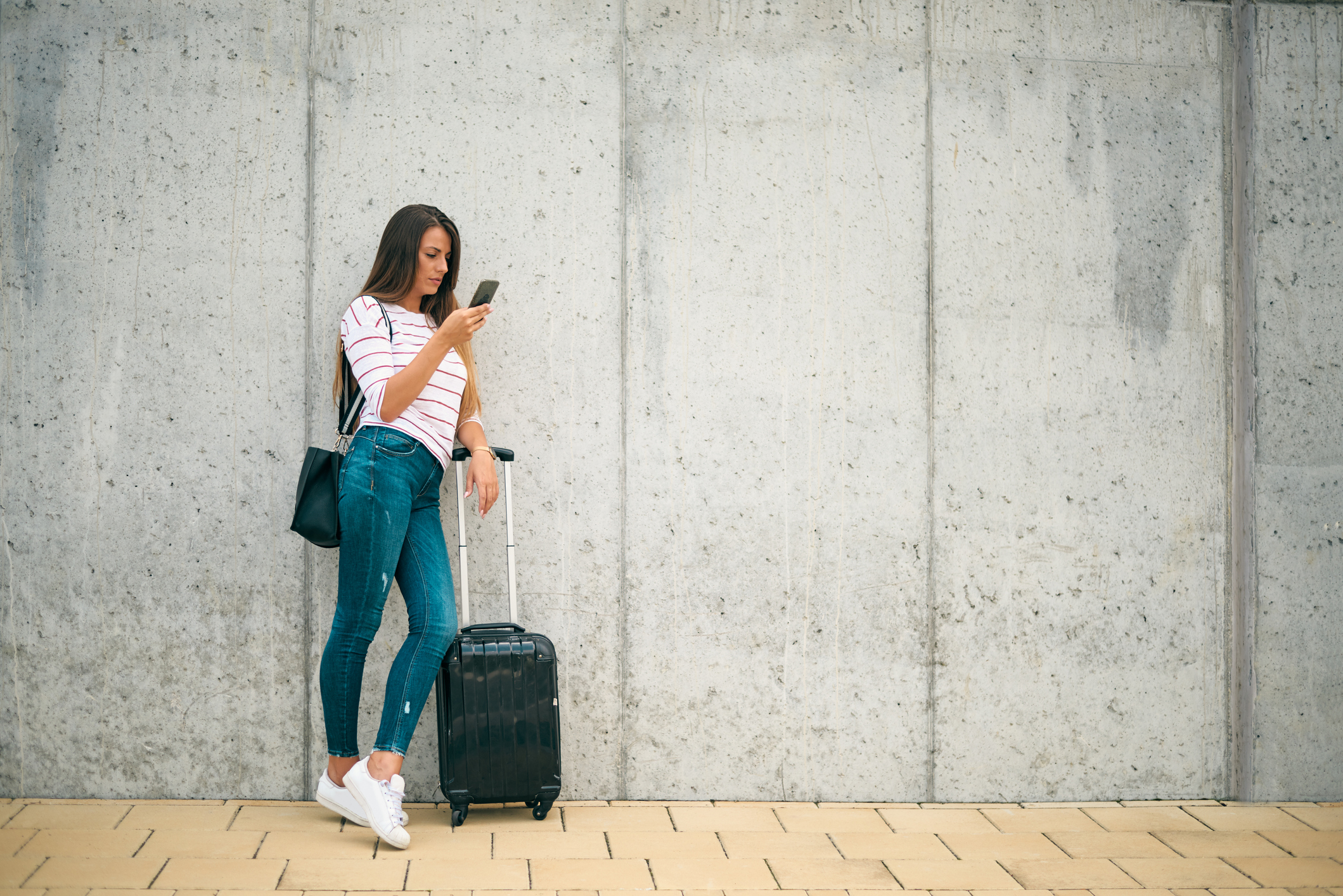 Mujer apoyada contra una pared, mirando el teléfono, con maleta