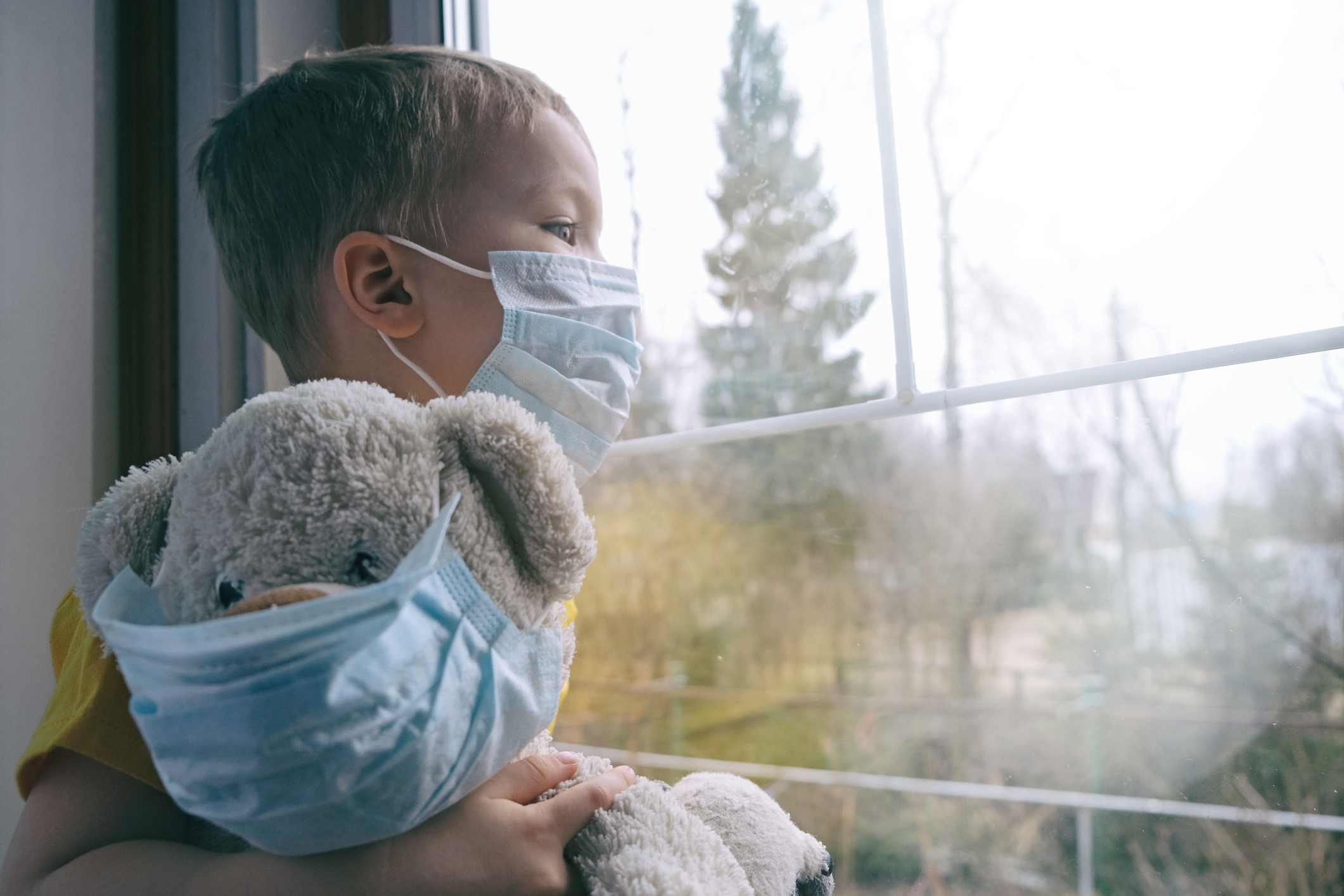 Child holding teddy bear, both wearing masks, looking out the window