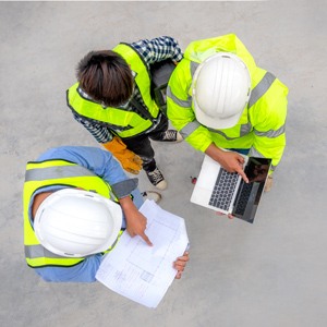 A top-down view shows three construction workers wearing hard hats and high-visibility safety vests collaborating on a job site. One worker points to printed building plans while another holds a laptop.